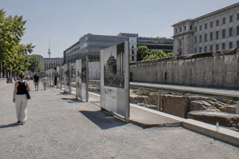 Long outdoor exhibition panels along a historic city street with a stone wall behind them