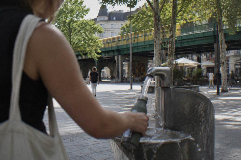 A person filling a water bottle at a Berlin drinking fountain on a sunny street