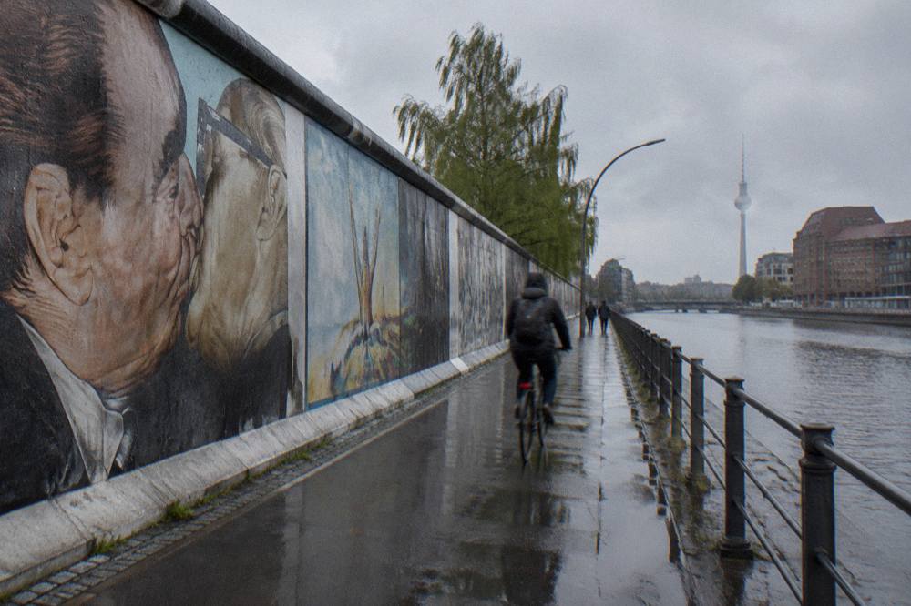 A long painted stretch of the former Berlin Wall standing beside a riverside path