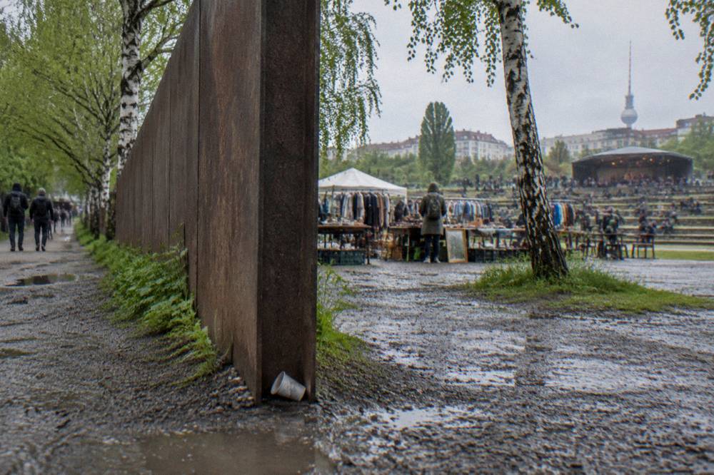 Section of a steel wall with a gap in it, set in a park with trees and a path beside it